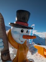 Ein bunter Schneemann mit Skibrille und Holzski vor Bergen und Zauchensee-Fahnen