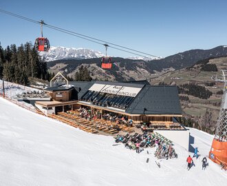 Skihütte mit Sonnenterrasse an einer Skipiste, darüber rote Gondeln und dahinter schneebedeckte Berge