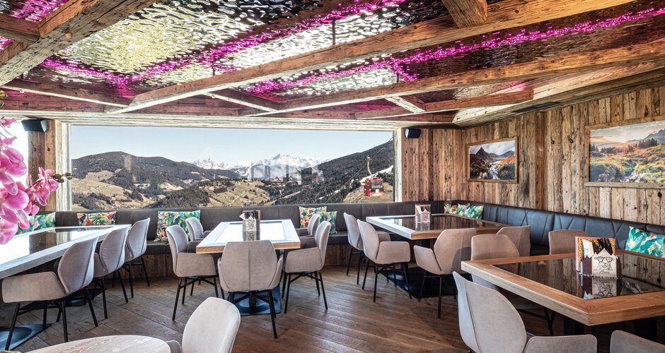 Rustic dining room with timber walls, tables, chairs, pink-lit ceiling and mountain view through a panoramic window