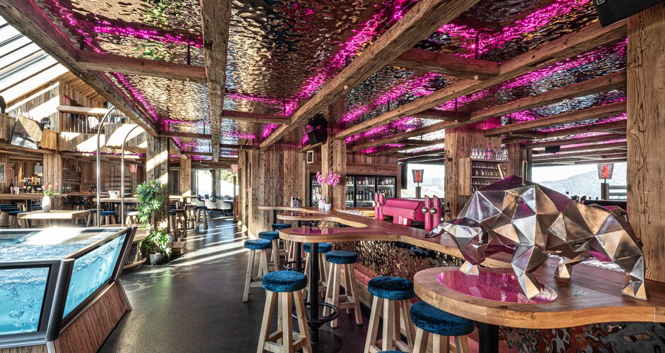 Rustic alpine bar with timber beams, pink-lit ceiling, stools, hot tub and metal bear sculpture