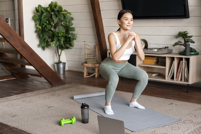 Fit woman doing squats on grey mat with dumbbell, bottle and laptop in living room interior