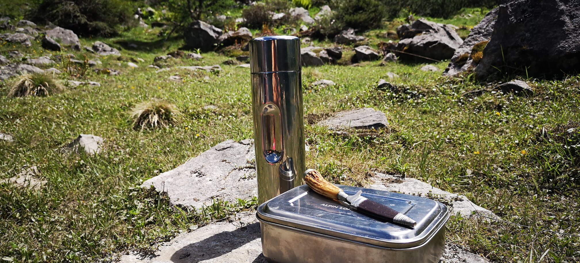 Metal lunchbox, knife and thermos bottle placed on rock in alpine meadow