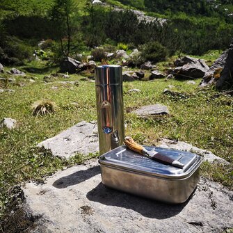 Metall-Jausenbox, Messer und Thermosflasche auf Fels in Almwiese platziert