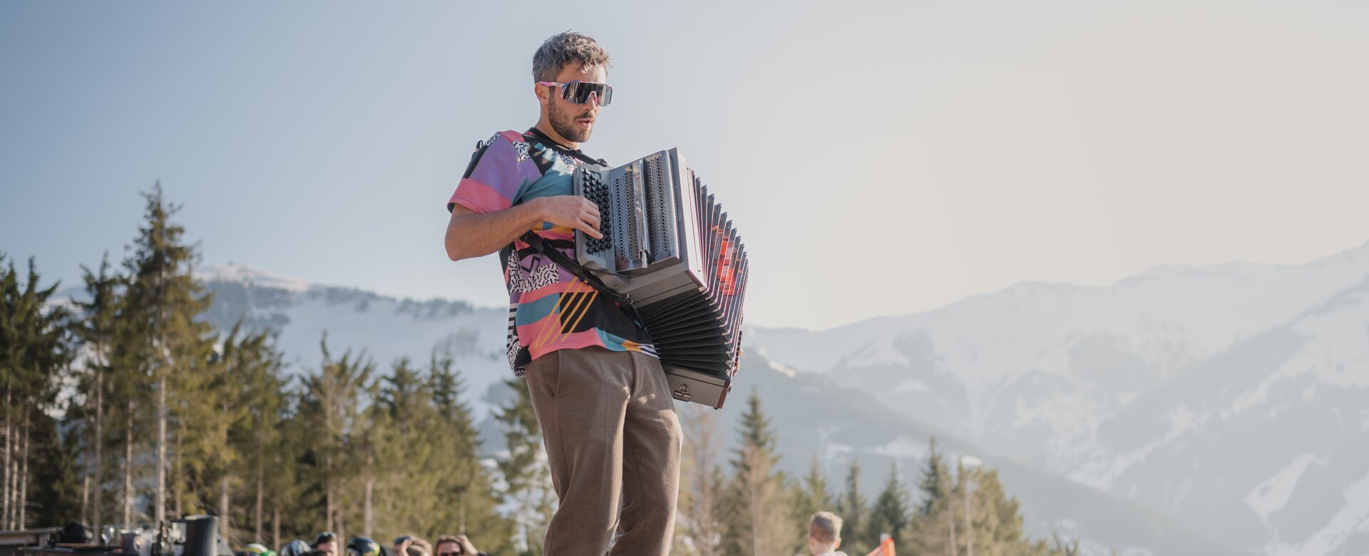 Mann mit Akkordeon spielt auf Tisch inmitten feiernder Gäste auf sonniger Terrasse vor Bergkulisse