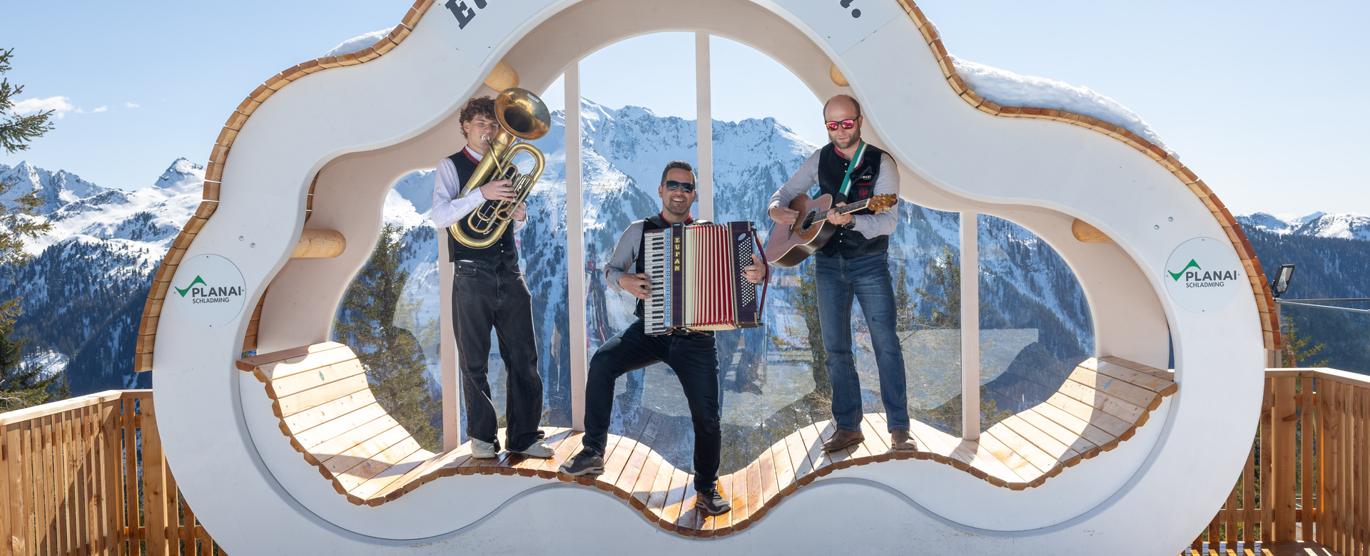 Drei Musikanten mit Tuba, Akkordeon und Gitarre spielen auf Holzplattform vor verschneiten Bergen | © Harald Steiner