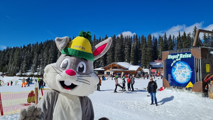 Hopsi is looking forward to the family festival | © Planai Hopsi the bunny mascot smiles at the camera and the family party takes place behind him | © Planai