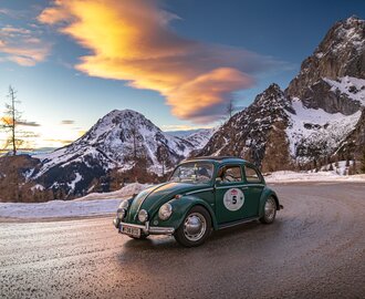 Ein grüner VW-Käfer fährt bei Sonnenuntergang auf verschneiter Passstraße vor Alpenpanorama | © Ondrey Kroutil