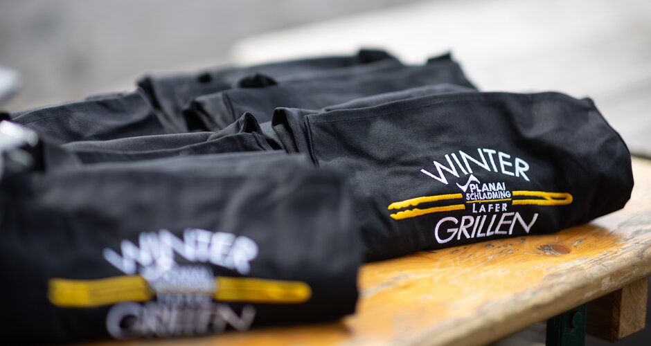 Mehrere schwarze Grillschürzen mit Logo „Wintergrillen Planai Schladming Lafer“ auf Tisch. | © Harald Steiner