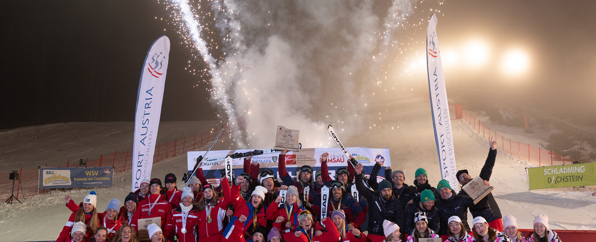 Große Siegergruppe posiert vor Feuerwerk und Bannern auf nächtlicher Skipiste | © Michael Simonlehner