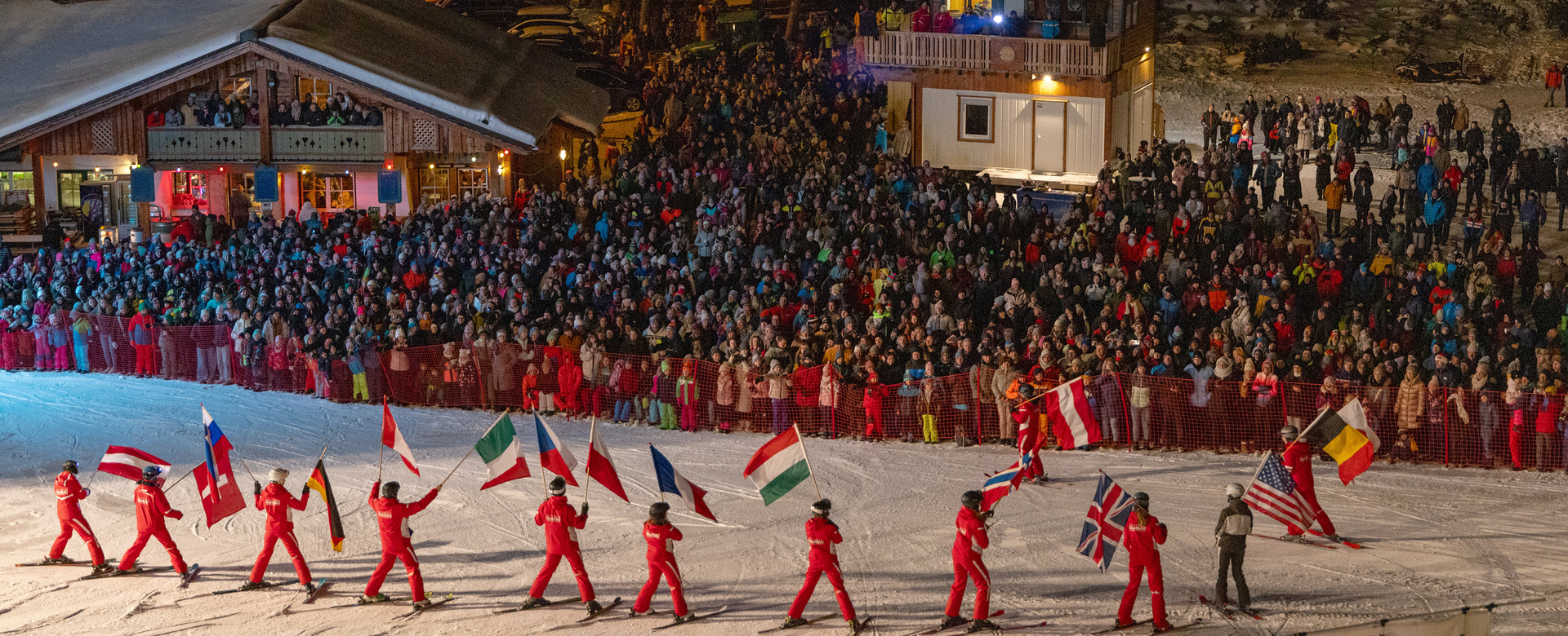 Skiers in red carry national flags on a slope at night in front of a large crowd | © Michael Simonlehner