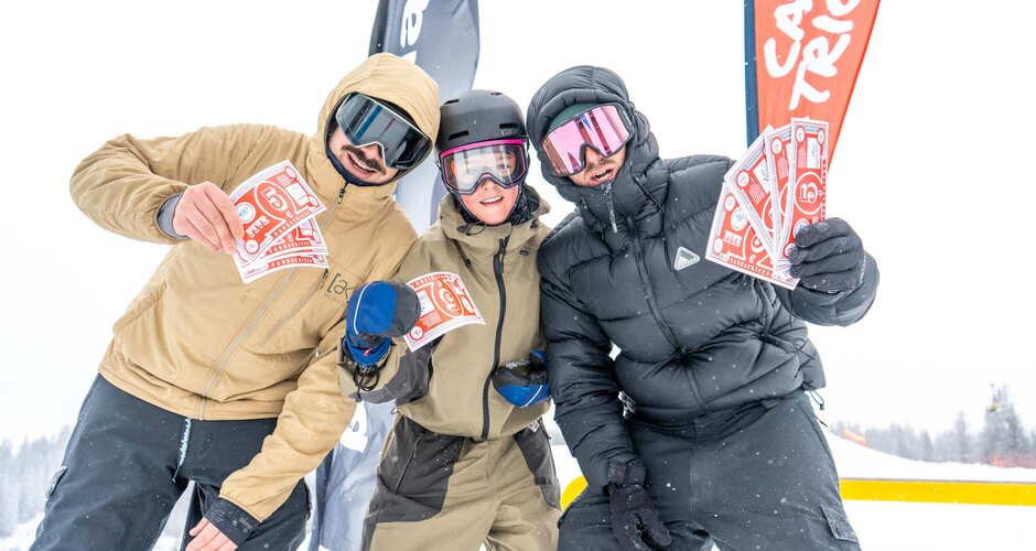 Drei Snowboarder halten bei Schneefall rote Gewinnscheine vor Fahnen im Superpark Planai in die Kamera | © Jordan Sitzwohl