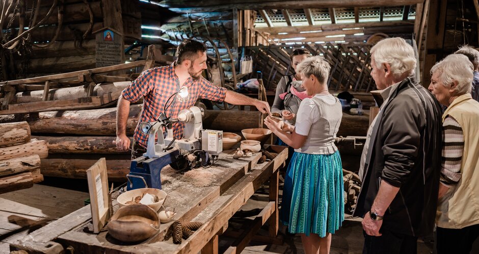 Handwerker zeigt Drechselarbeiten aus Holz, Besuchergruppe schaut interessiert bei traditionellem Handwerk in alter Werkstatt zu. | © Matthias Fritzenwallner