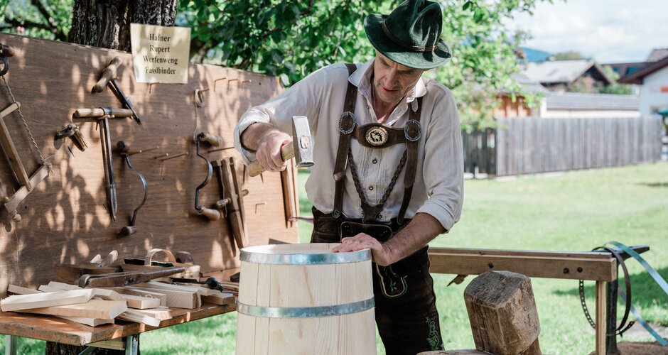 Fassbinder in Lederhose arbeitet mit Hammer an Holzfass, umgeben von traditionellem Werkzeug bei Handwerksvorführung. | © Matthias Fritzenwallner