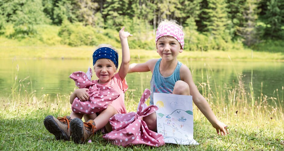Two smiling kids with headbands sit by the lake, holding picnic bags and a happy, self-drawn picture with sun and mountains. | © Matthias Fritzenwallner