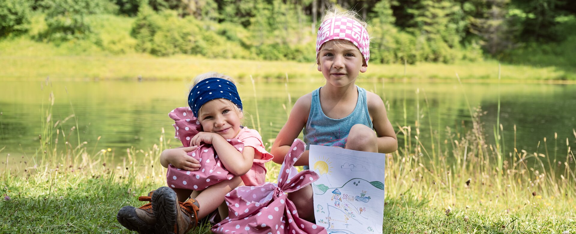 Zwei Kinder sitzen am See beim Event See.Musik, mit Picknickbeuteln und einem selbstgemalten Bild mit Sonne und Bergen. | © Matthias Fritzenwallner