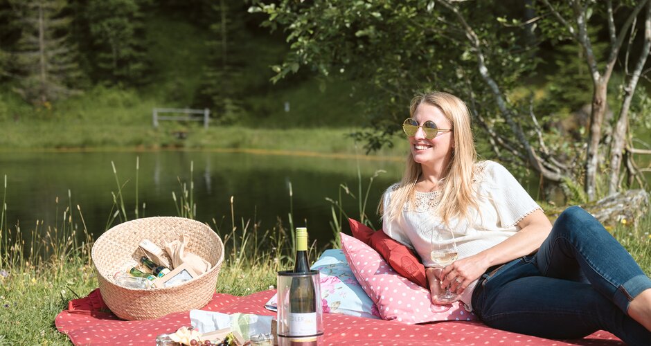 Frau liegt auf gepunkteter Decke am See, mit Weinglas, Picknickkorb und Weinflasche beim Event See.Musik in der Sonne | © Matthias Fritzenwallner