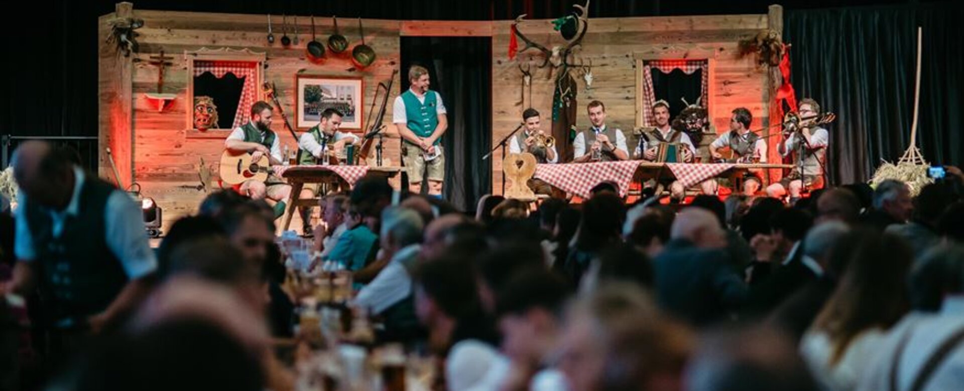 Musiker in Tracht auf Bühne beim Gstanzlsingen in Altenmarkt, davor dicht besetzte Tische mit Publikum in festlicher Stimmung. | © TVB Altenmarkt-Zauchensee