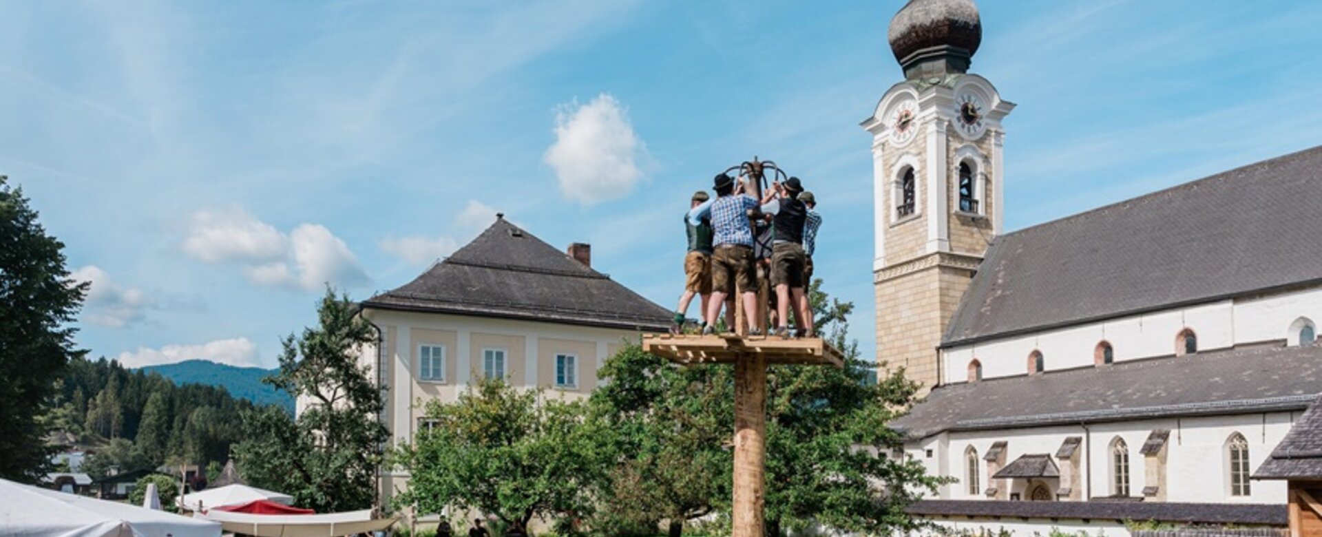 Männer in Tracht stehen auf einem Kletterbaum beim Fest im Pfarrhof Altenmarkt, Zuschauer sitzen auf Bierbänken im Grünen. | © Matthias Fritzenwallner