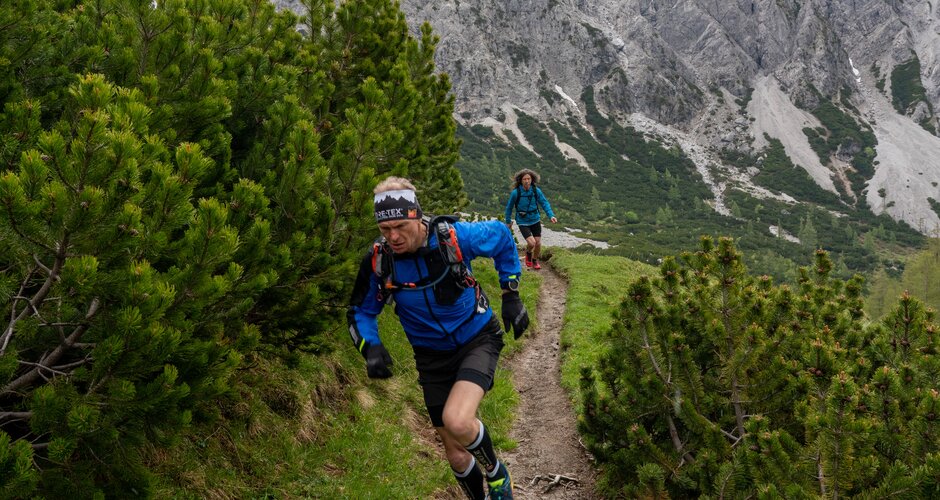 Ein Trailrunner läuft einen schmalen, steilen Pfad durch Latschenkiefern mit Blick auf schneebedeckte Felsgipfel im Hintergrund. | © ©CoenWeesjes_www.filzmoos.at