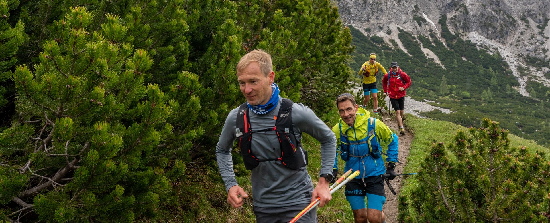 Eine Gruppe Trailrunner läuft auf einem schmalen Bergpfad durch Latschenkiefern mit Blick auf schneebedeckte Felsgipfel. | © ©CoenWeesjes_www.filzmoos.at