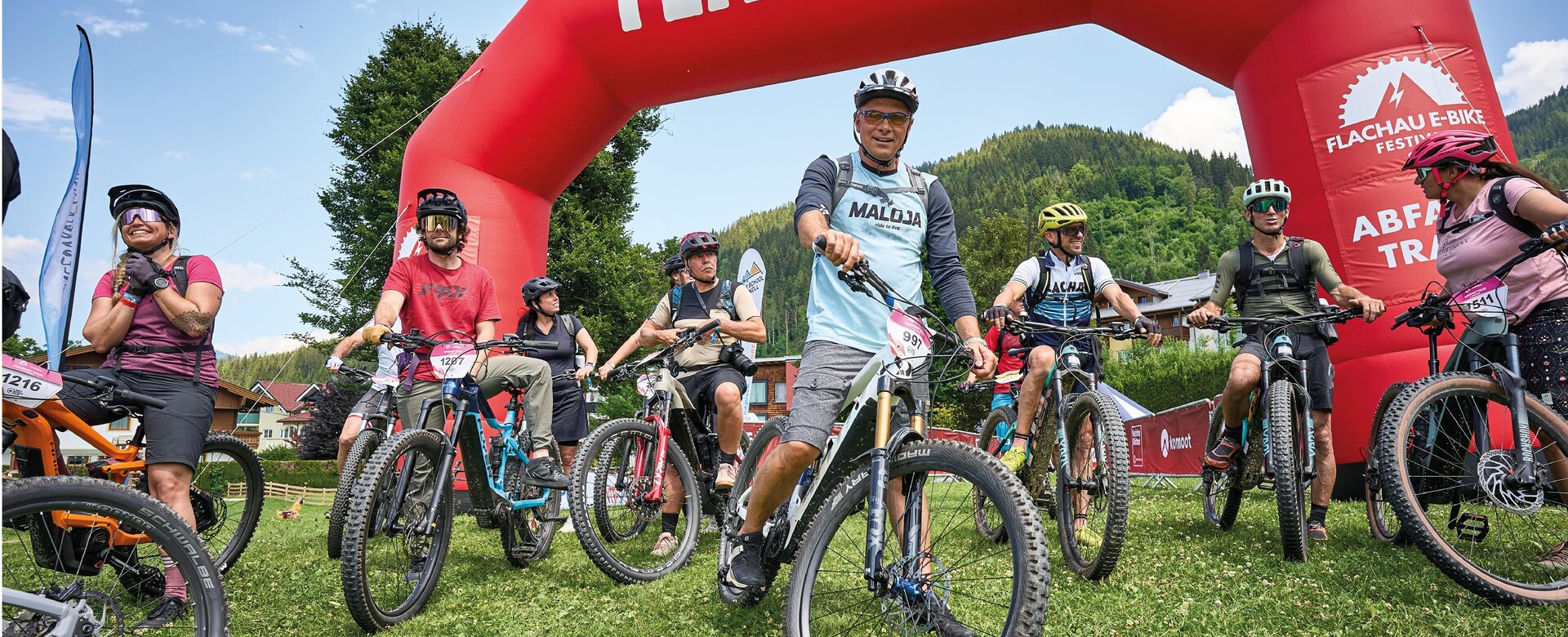 Mehrere E-Biker beim Start unter rotem Flachau-Bogen vor grüner Bergkulisse. | © Flachau Tourismus