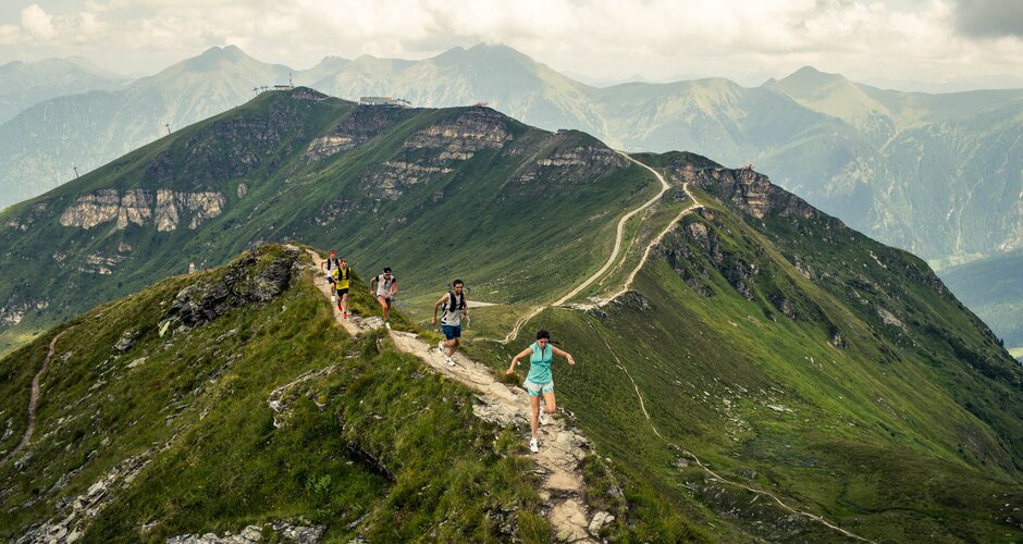 Mehrere Trailrunner:innen laufen über einen schmalen Berggrat mit Blick auf grüne Alpengipfel und wolkenverhangenen Himmel | © Gasteinertal Tourismus GmbH, Michael Mueller