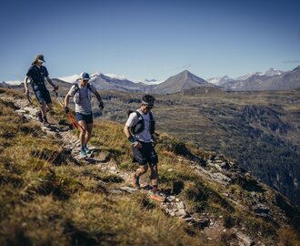 Drei Trailrunner wandern über einen felsigen Bergpfad mit Blick auf grüne Täler und die schneebedeckten Hohen Tauern | © Stefan Schaaf PEAK ART IMAGES