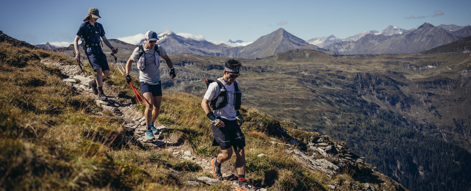Drei Trailrunner wandern über einen felsigen Bergpfad mit Blick auf grüne Täler und die schneebedeckten Hohen Tauern | © Stefan Schaaf PEAK ART IMAGES