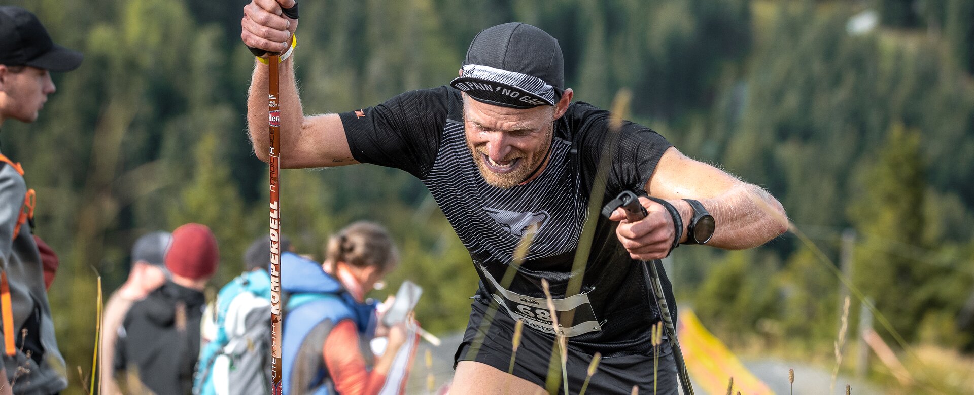 Ein Trailrunner kämpft sich mit Stöcken den steilen Hang hinauf, im Hintergrund Zuschauer und Berglandschaft. | © Beinhoat