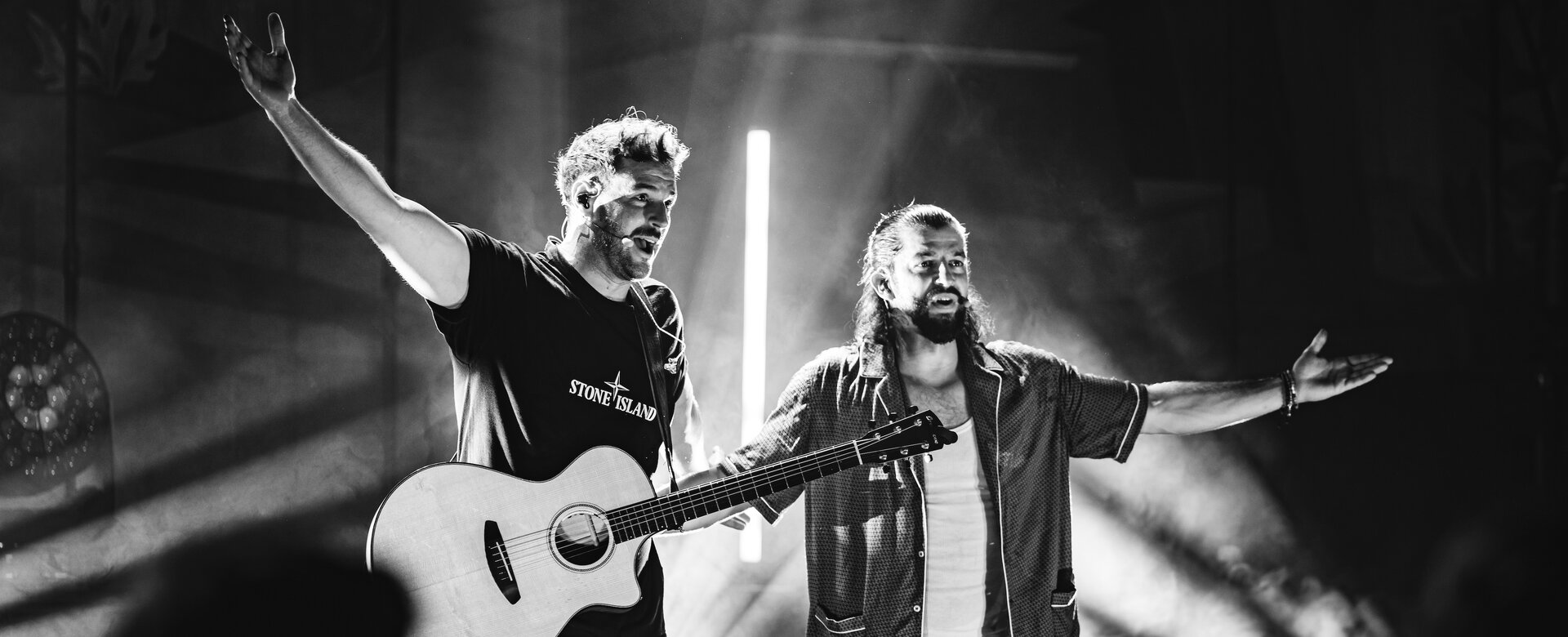 Two musicians on stage, one with a guitar, both with arms wide open under dramatic stage lighting. | © Pascal Riesingerm