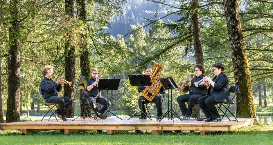 Ein Bläserensemble spielt ein Konzert auf einer Waldbühne, umgeben von Bäumen und Sommerlicht in Gastein | © Gasteinertal Tourismus GmbH, Simon Hutter