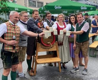 Gruppe in Tracht beim Bieranstich mit Krügen auf Straße vor Kirche und Bergen.