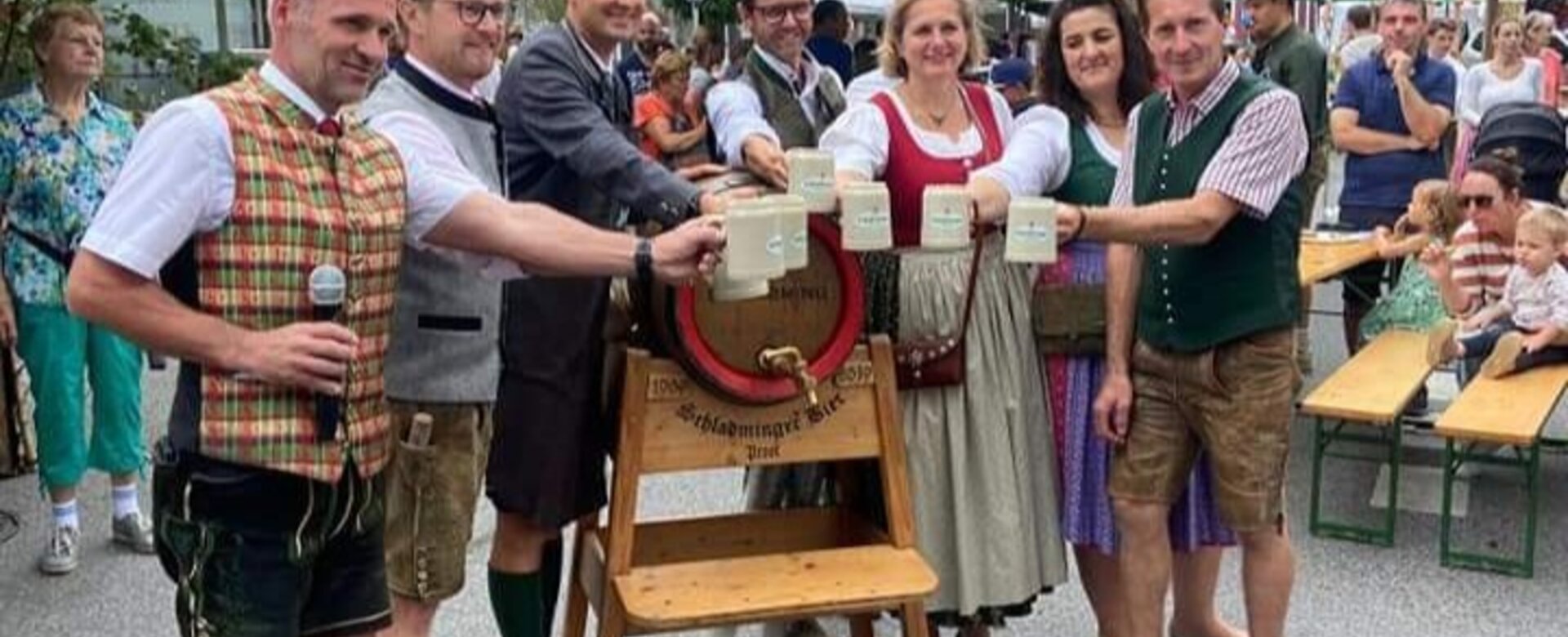 Gruppe in Tracht beim Bieranstich mit Krügen auf Straße vor Kirche und Bergen.