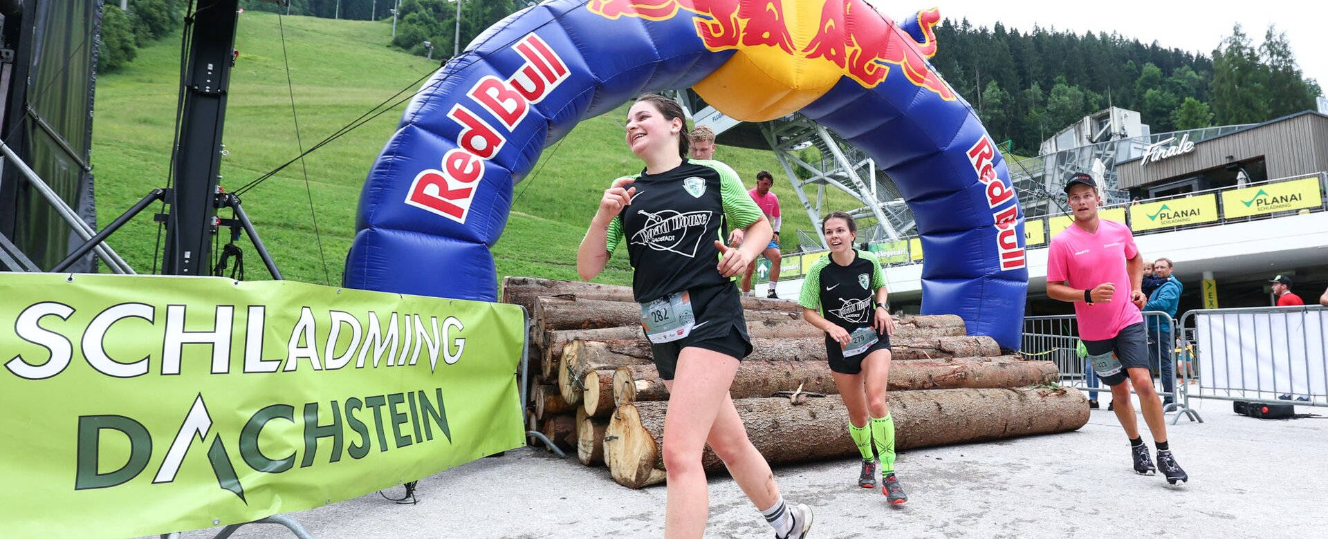 Mehrere Läufer überwinden ein Hindernis aus Baumstämmen unter dem Red-Bull-Bogen beim Tauern Circle in Schladming. | © David Geieregger