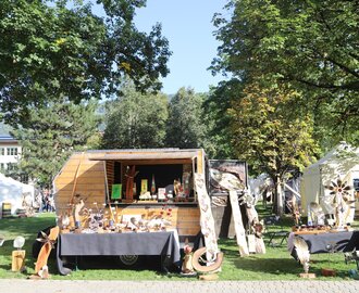 Marktstand mit Holzkunst, Metall-Deko und Handwerk im Park, sonniger Tag, weiße Zelte im Hintergrund | © JOSalzburg