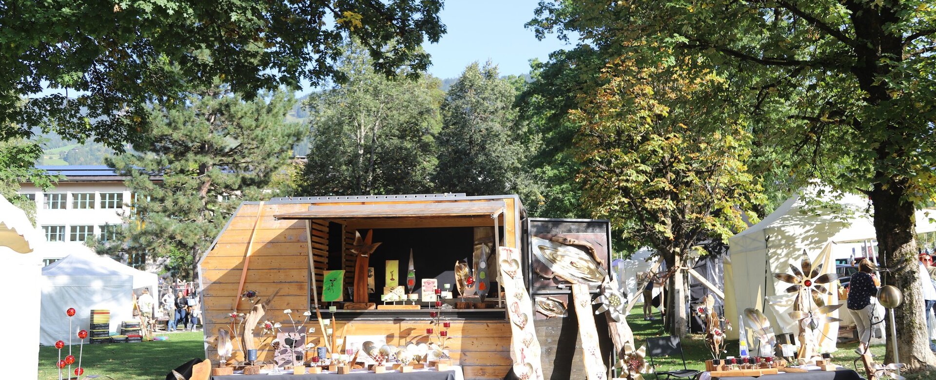 Marktstand mit Holzkunst, Metall-Deko und Handwerk im Park, sonniger Tag, weiße Zelte im Hintergrund | © JOSalzburg