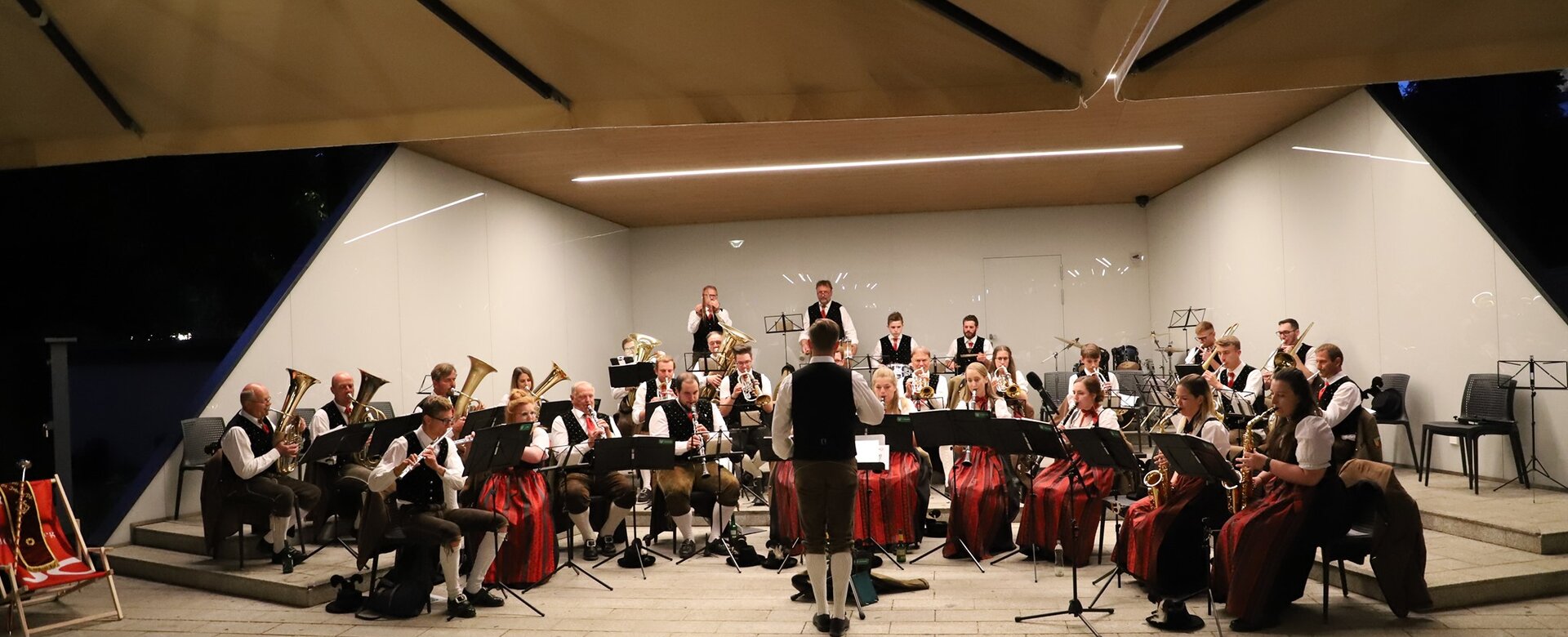 Brass band in traditional costume at evening town concert on open-air stage, conductor in front | © Bürgermusik