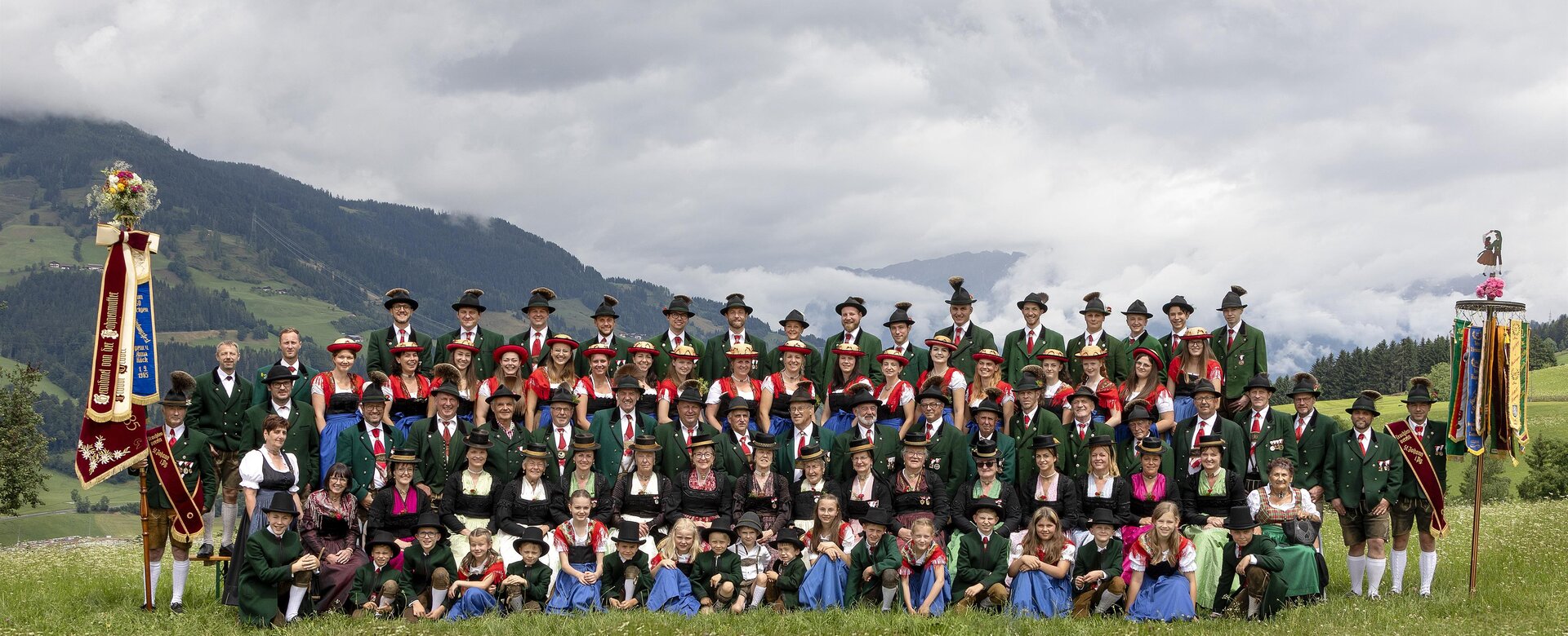 Große Gruppe in Tracht mit zwei Vereinsfahnen auf Almwiese, Männer in Grün, Frauen in Dirndl, Kinder vorn | © Trachtenverein