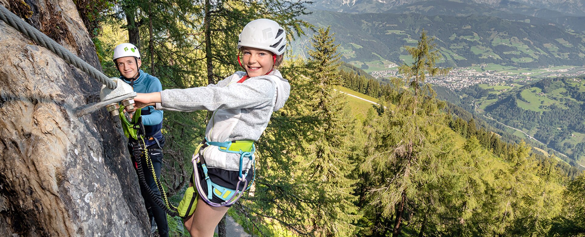 Two kids with climbing gear on rock wall with view over green alpine valley | © Christoph Huber