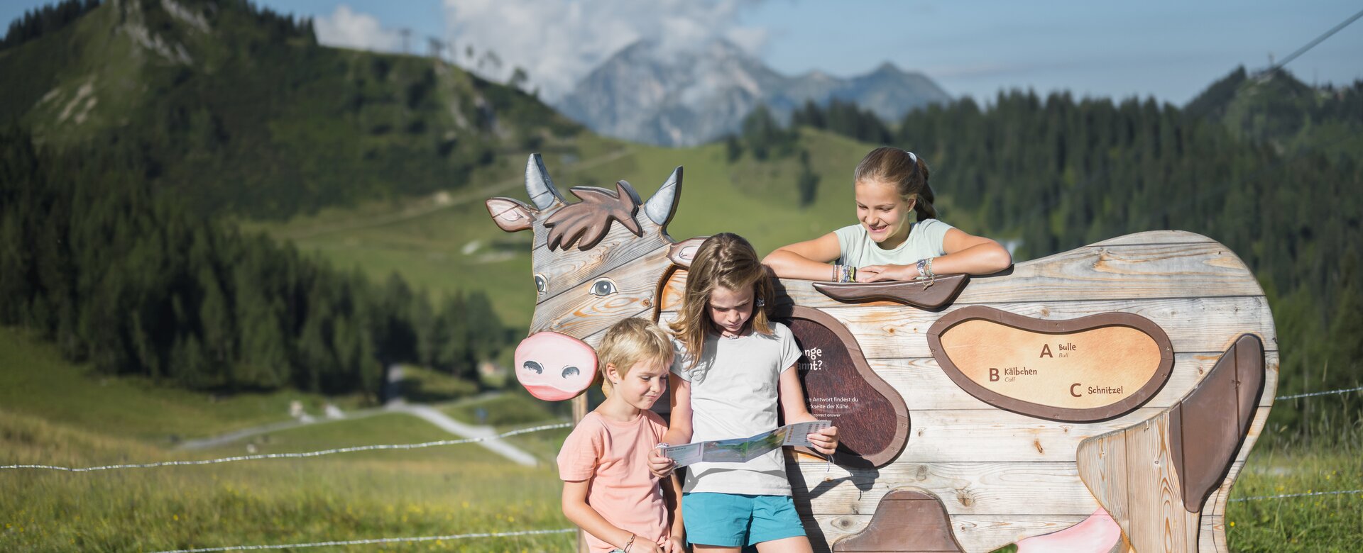 Three kids at wooden cow station on KUHbidu trail in green alpine landscape | © Christian Schartner