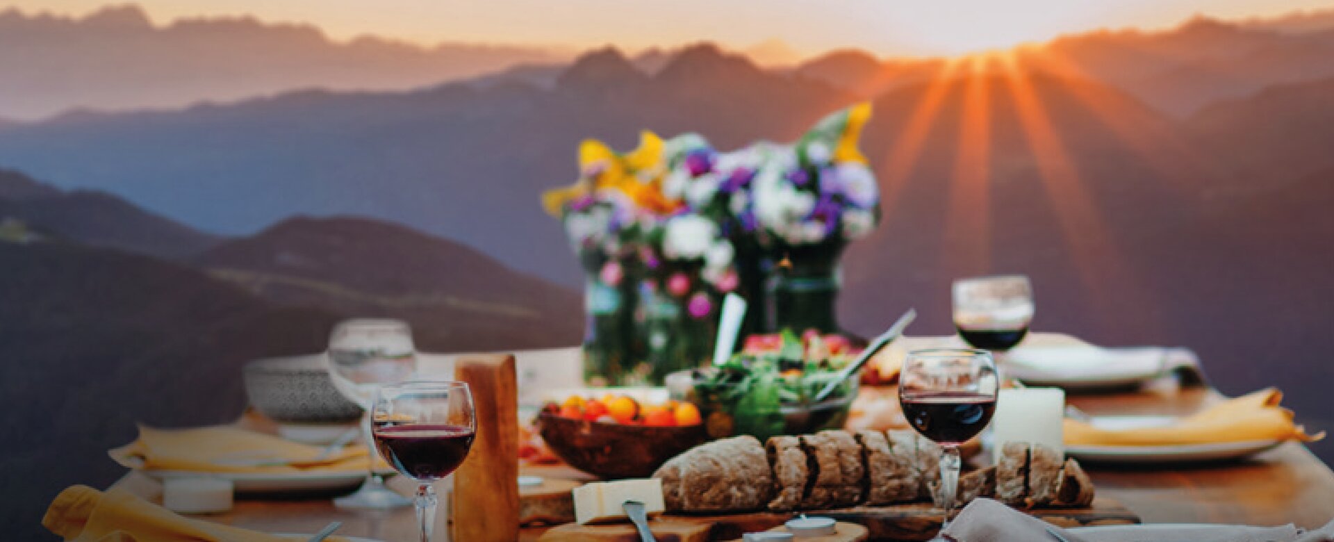 Holztisch mit Wein, Brot, Käse und Blumen beim Abendessen in den Bergen bei Sonnenuntergang