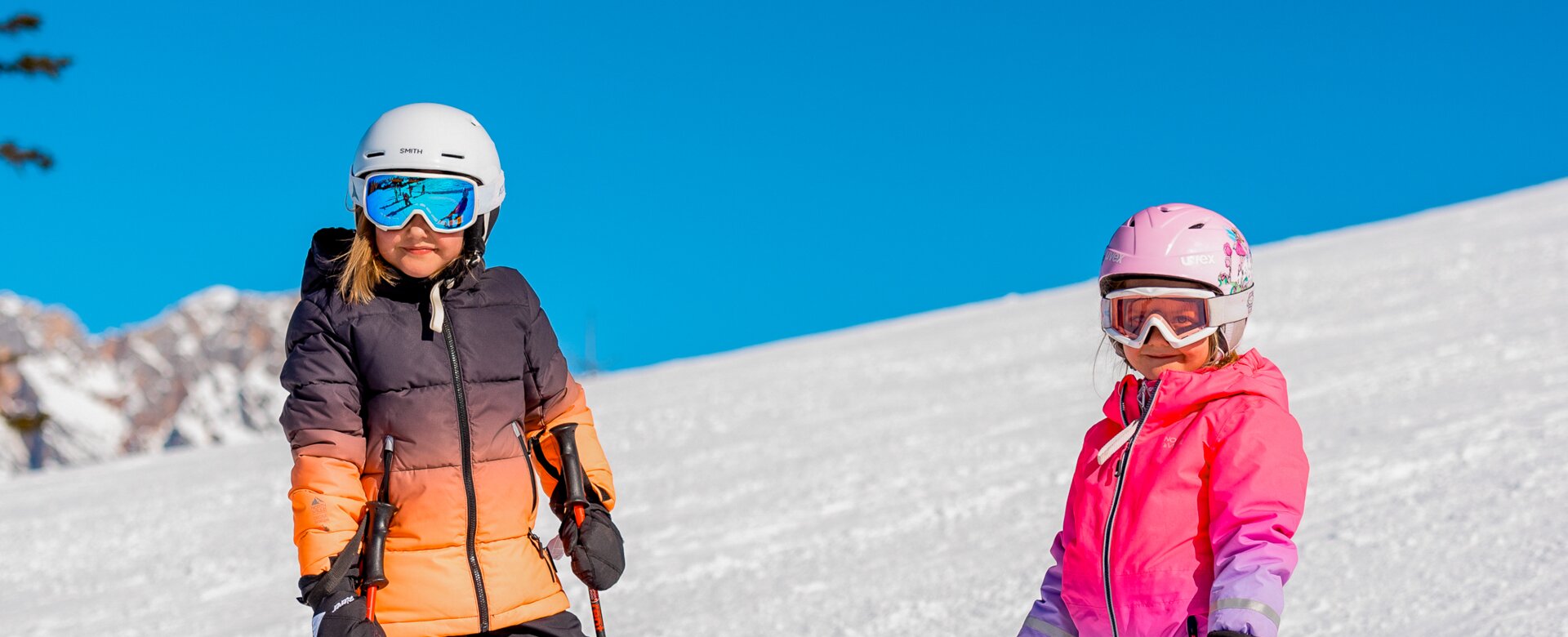 Zwei Kinder im Skianzug und Helm stehen auf einer sonnigen Skipiste. | © Hochkönig Tourimus GmbH