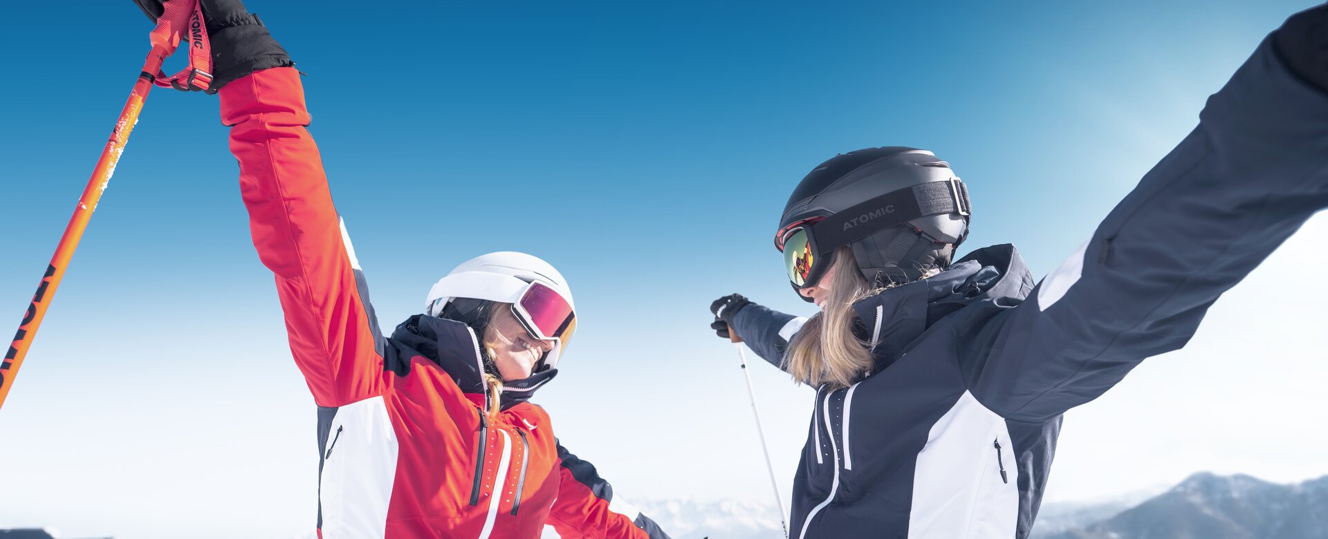 Zwei Frauen in moderner Skibekleidung, eine in einer roten Jacke mit weißem Helm und Skibrille, die andere in einer schwarzen Jacke mit schwarzem Helm, stehen auf einem schneebedeckten Berg und strecken fröhlich die Arme aus. Im Hintergrund ist ein strahlend blauer Himmel sowie ein Panorama mit Bergen und einem Skilift zu sehen.