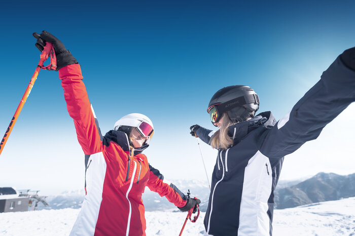 Feeling joy while learning to ski Zwei Frauen in moderner Skibekleidung, eine in einer roten Jacke mit weißem Helm und Skibrille, die andere in einer schwarzen Jacke mit schwarzem Helm, stehen auf einem schneebedeckten Berg und strecken fröhlich die Arme aus. Im Hintergrund ist ein strahlend blauer Himmel sowie ein Panorama mit Bergen und einem Skilift zu sehen.