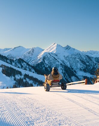 Person fährt Schneekart auf frisch präparierter Piste mit Blick auf verschneite Berge bei Sonnenschein. | © Dorfgasteiner Bergbahnen AG