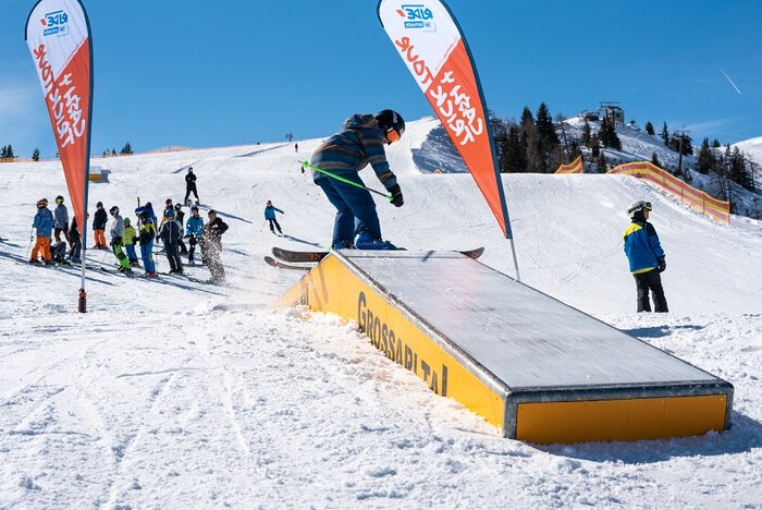 Teen slides box during Cash4Tricks session in Grossarl Young freestyler slides yellow box with Cash4Tricks flags, crowd of spectators watching on sunny snowy slope in background.