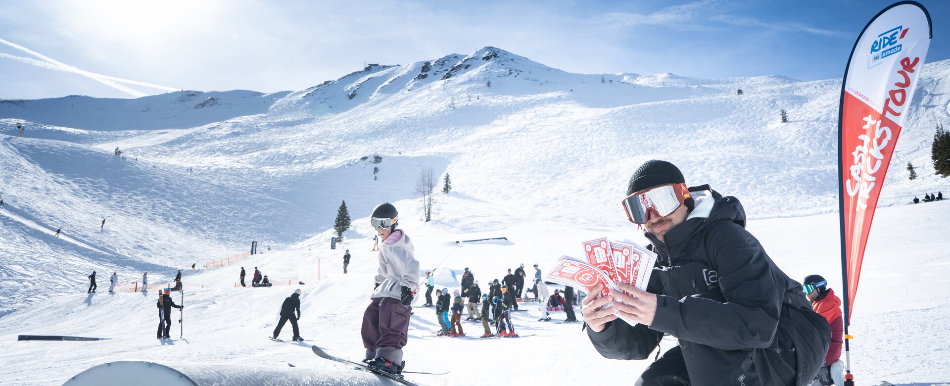 Ein Judge kniet im Schnee mit roten Cash4Tricks-Gutscheinen, viele Zuschauer bei strahlendem Sonnenschein.