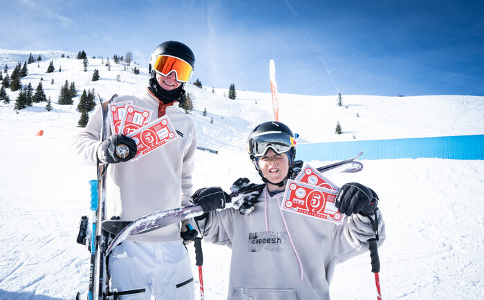 Two freeskiers show off their prizes at Cash4Tricks Two young freeskiers hold red Cash4Tricks vouchers, posing in snowpark scenery with skis and blue park tube behind them.