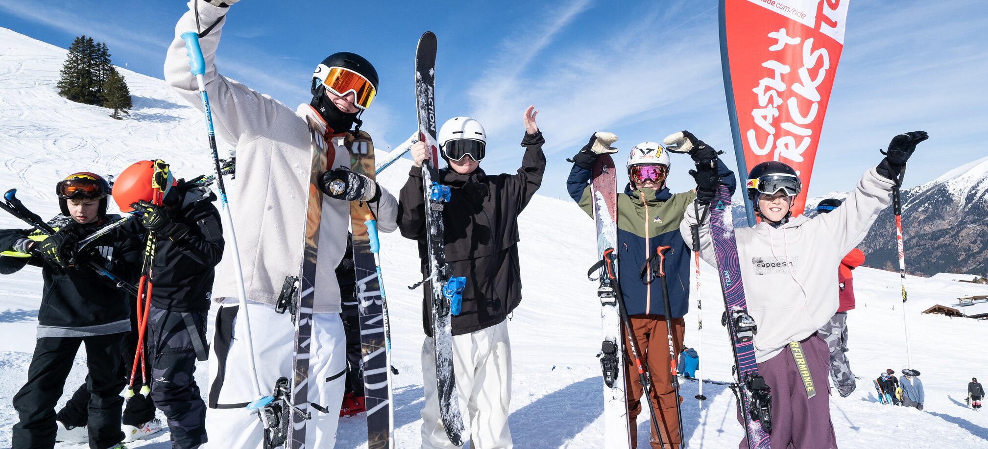 Four young freeskiers cheer in front of a Cash4Tricks flag, holding skis in the snow with blue sky and mountains behind them.