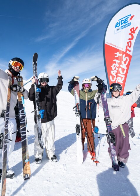 Vier junge Freeskier jubeln vor einer Cash4Tricks-Flagge im Schnee, Ski in der Hand, blauer Himmel und Berge im Hintergrund.